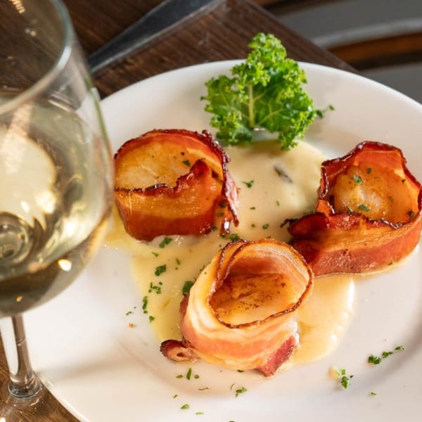 Three bacon-wrapped scallops on a white plate with creamy sauce, garnished with kale and parsley, next to a wine glass.