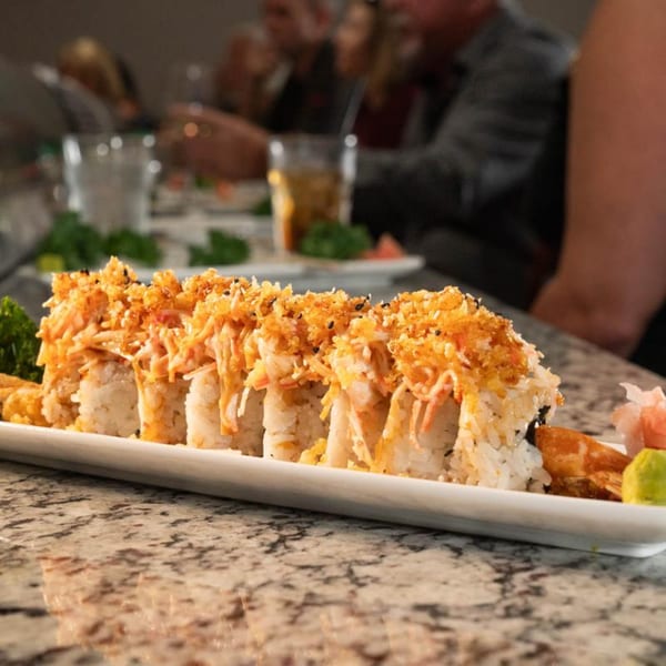 A sushi roll with crab and crispy topping on a white plate, with diners in the background.