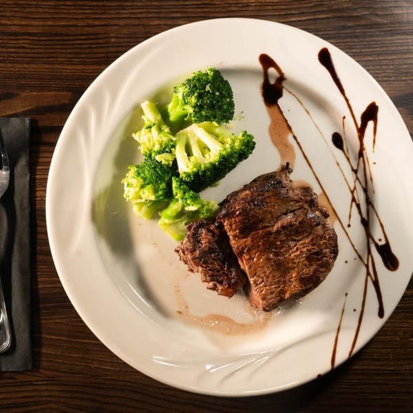 Grilled steak with a side of broccoli florets, drizzled with balsamic glaze on a white plate.