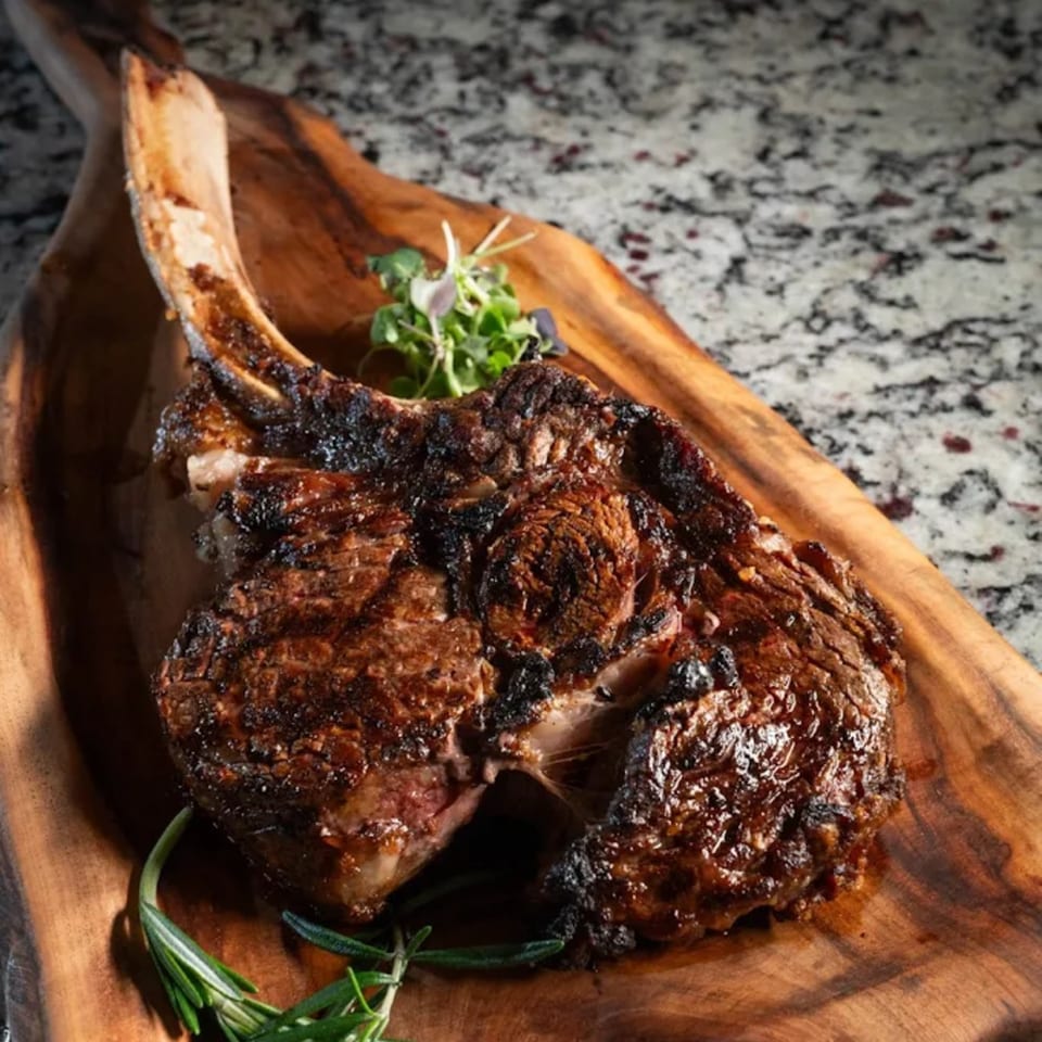 Grilled tomahawk steak with fresh herbs on a wooden board at Birkin's Steakhouse.