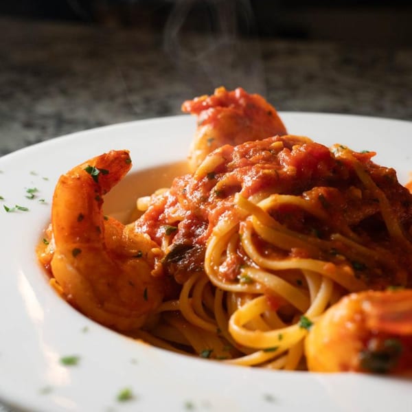 Steaming shrimp scampi with linguine pasta and red sauce, garnished with parsley in a white bowl.