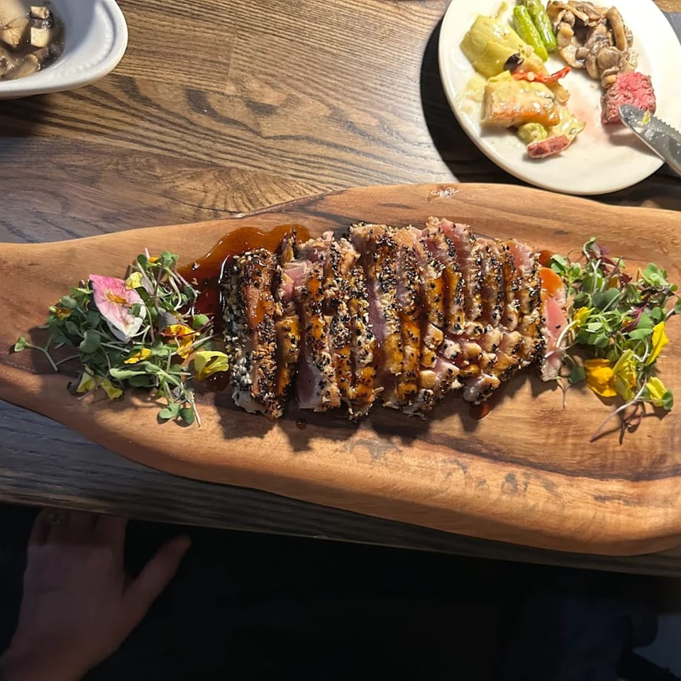 Seared tuna with sesame seeds and sauce, garnished with microgreens and edible flowers on a wooden board.